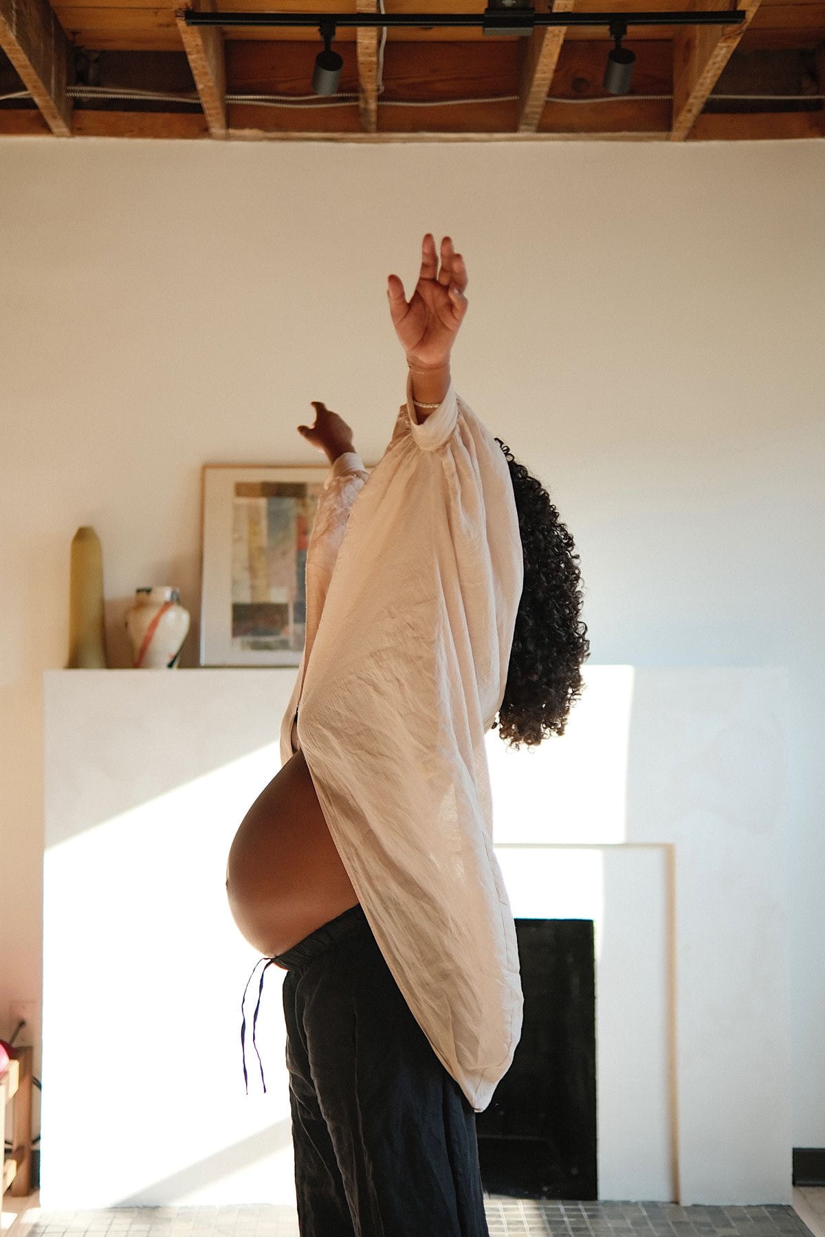 Pregnant Black mother with arms raised in a sun-filled room, draped in linen
